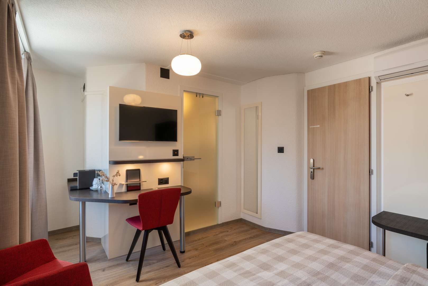 Habitación de hotel con cama, escritorio con silla, televisor montado en la pared y una puerta.