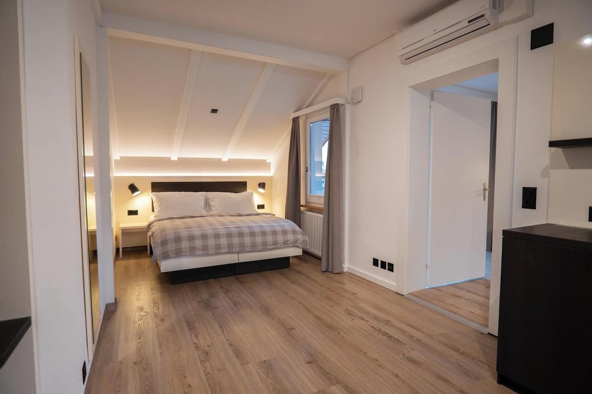 A modern bedroom with a large bed, bedside lamps, a window with curtains, and a wooden floor.