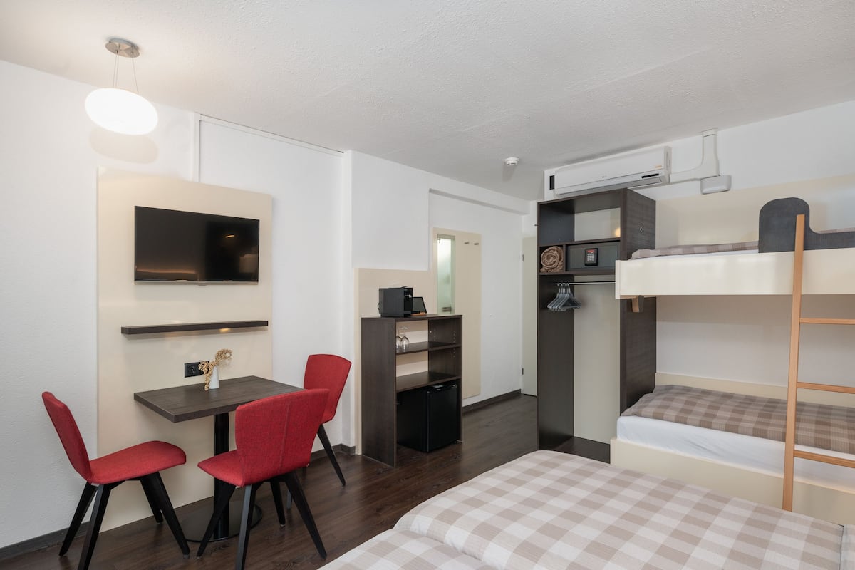 A compact room with a bunk bed, a desk with two red chairs, a small TV, and a kitchenette.
