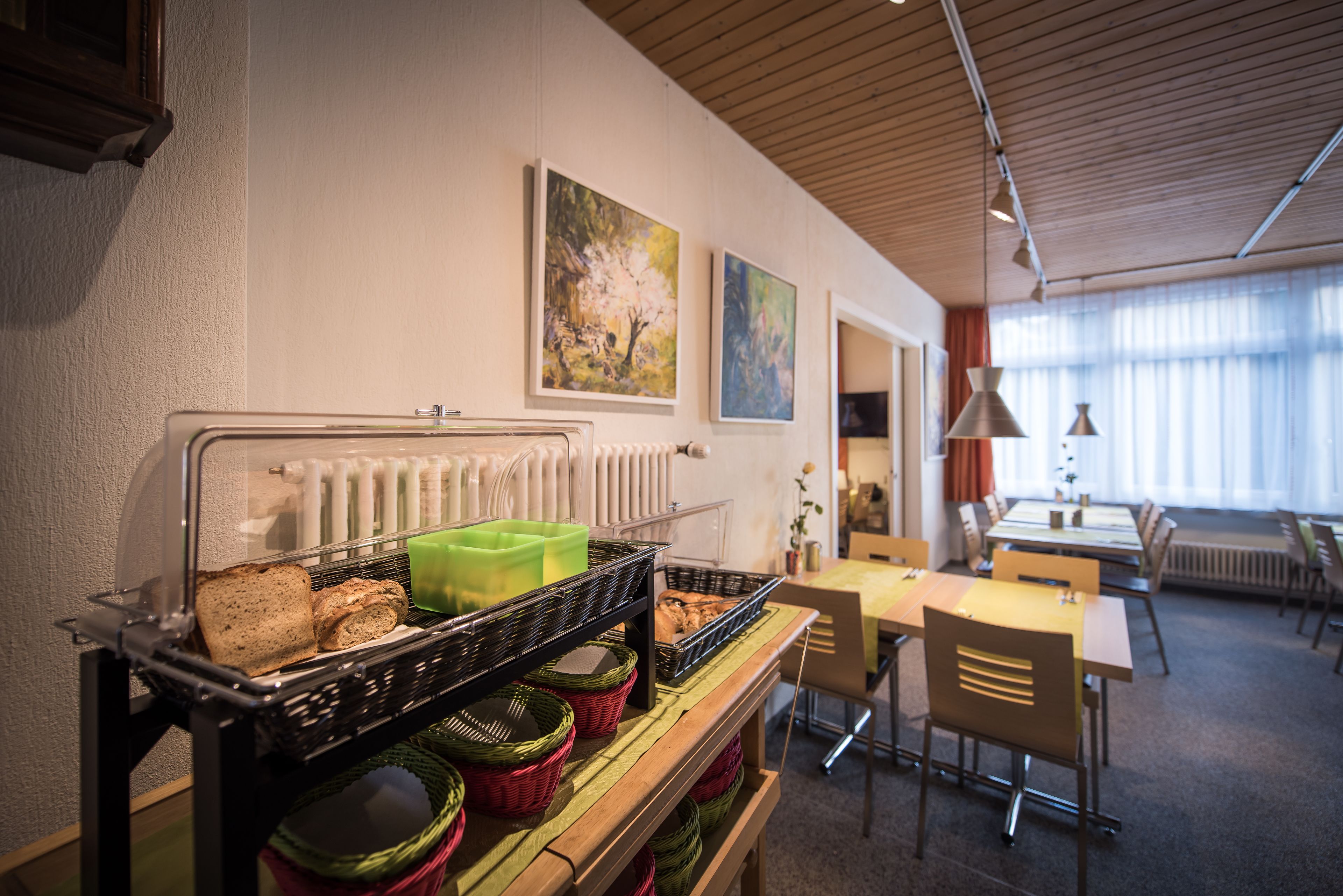 Ein Frühstücksbuffet mit Brot, Gebäck und Korbflexweben, ein Essbereich mit Tischen und Stühlen sowie zwei Bilder an der Wand.
