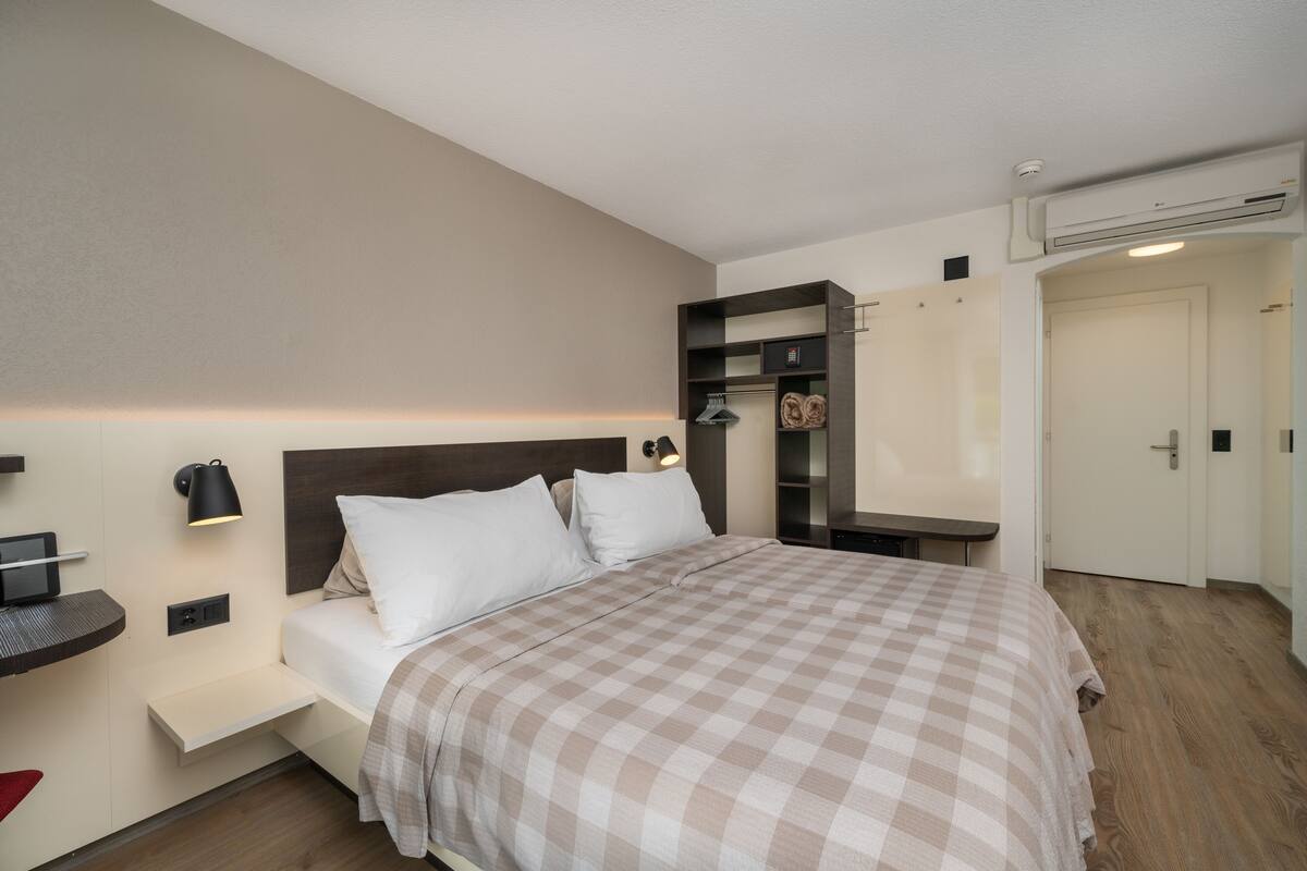 A modern hotel room with a bed, bedside table, wall-mounted lamp, and built-in wardrobe.