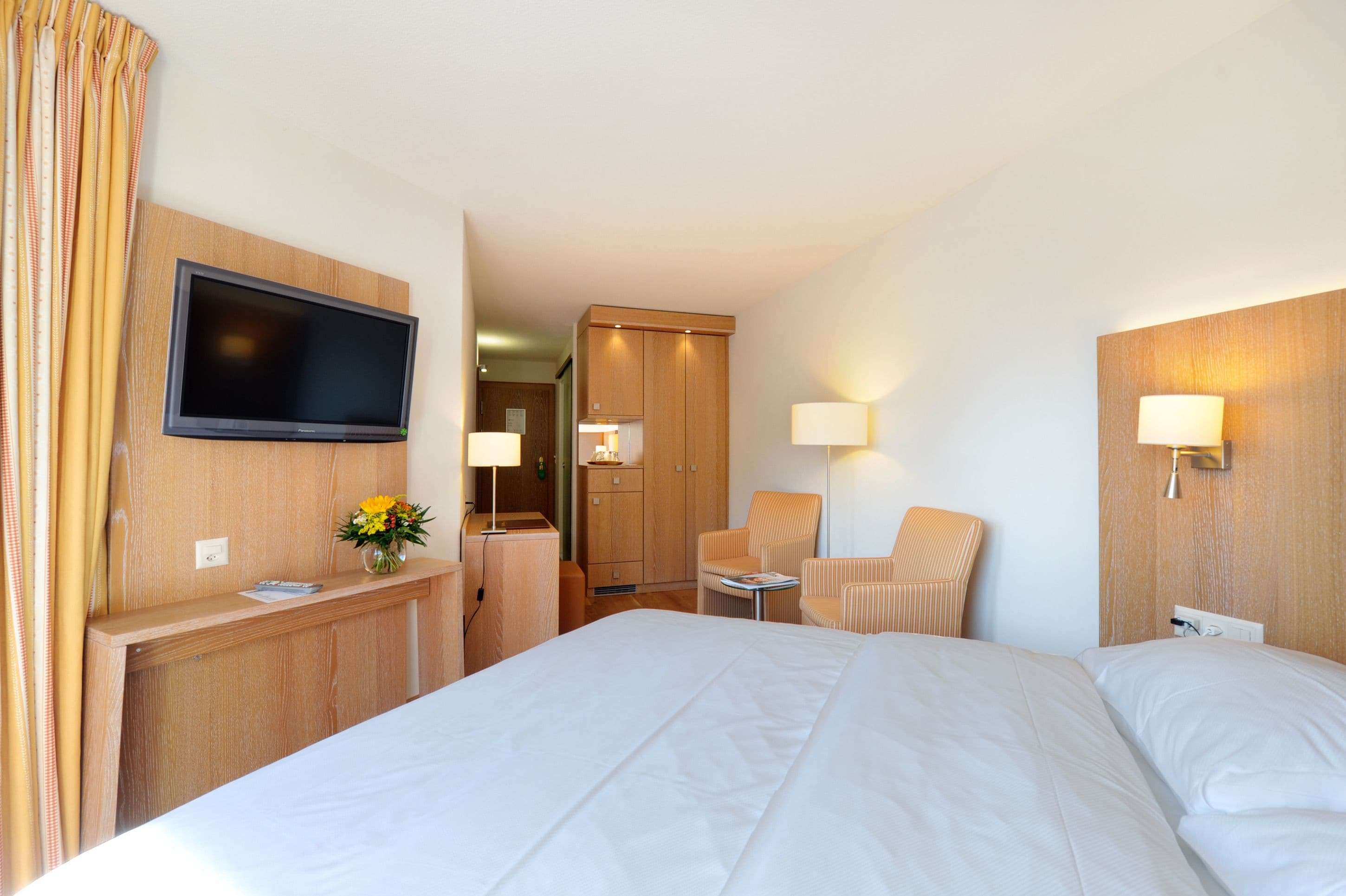 Een hotelkamer met een groot bed, een bureau met een flatscreen televisie, twee fauteuils, een bijzettafeltje en een houten garderobe.