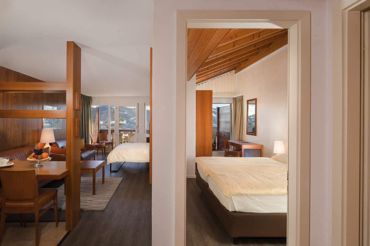 A hotel room with a wooden ceiling, a bed, a desk with a lamp, a chair, and a view of the mountains through large windows.