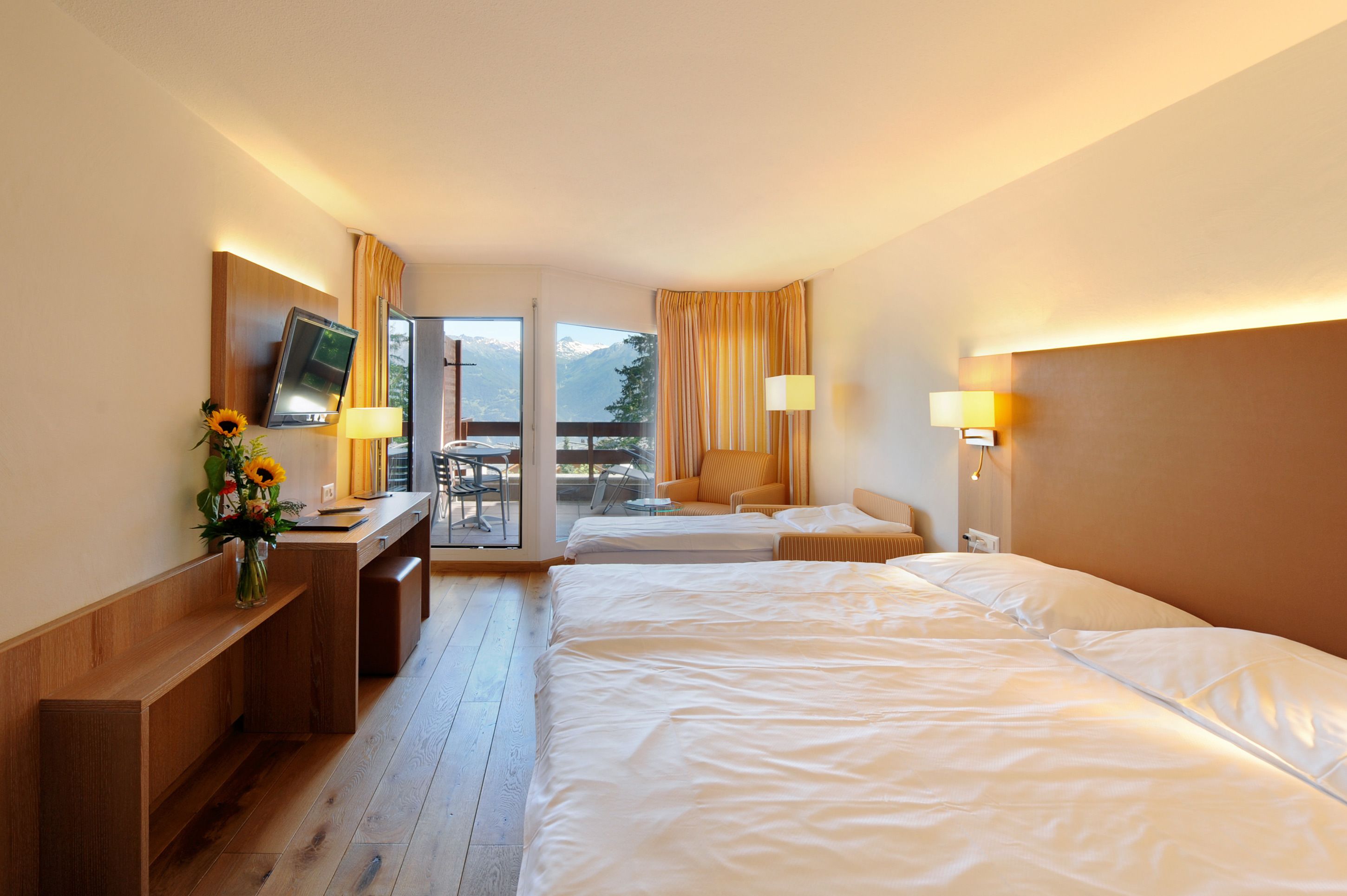 Habitación de hotel con dos camas, un escritorio y televisión, y vistas a montañas a través de la ventana.