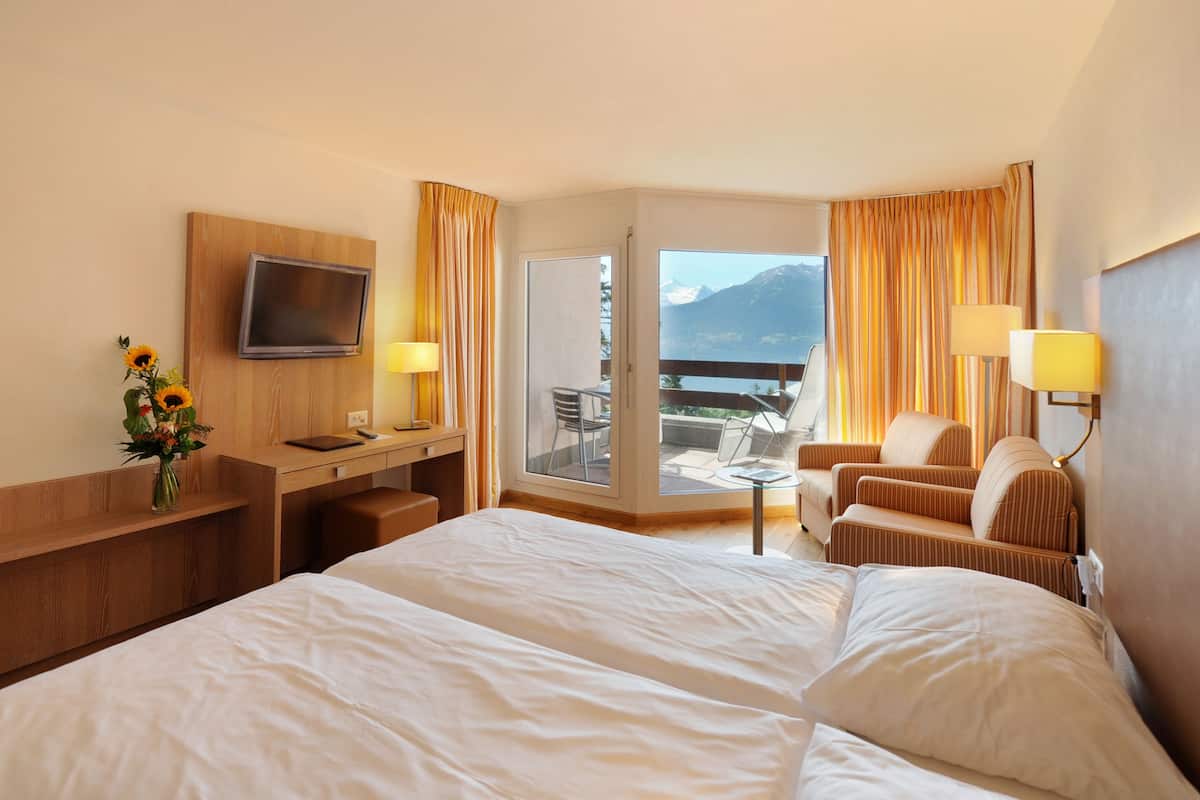 A hotel room with a large bed, a desk with a chair, a TV mounted on the wall, and a balcony with a chair and table.