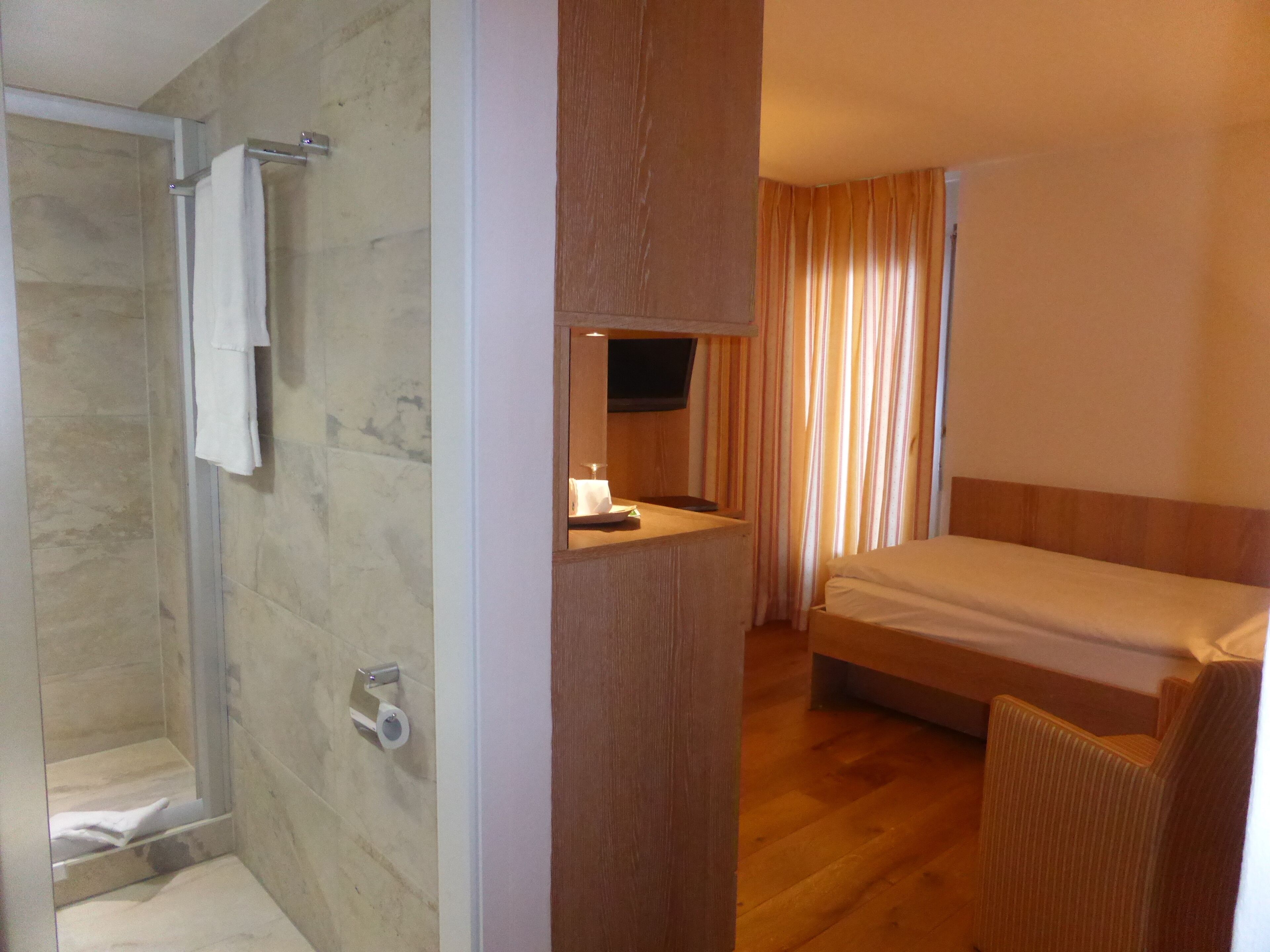 A hotel room with a shower, a bed, a wooden panel, and a television.
