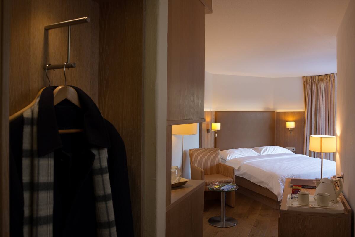 A hotel room with a bed, bedside lamps, a chair, a small table with a tray, and a hanging coat rack.