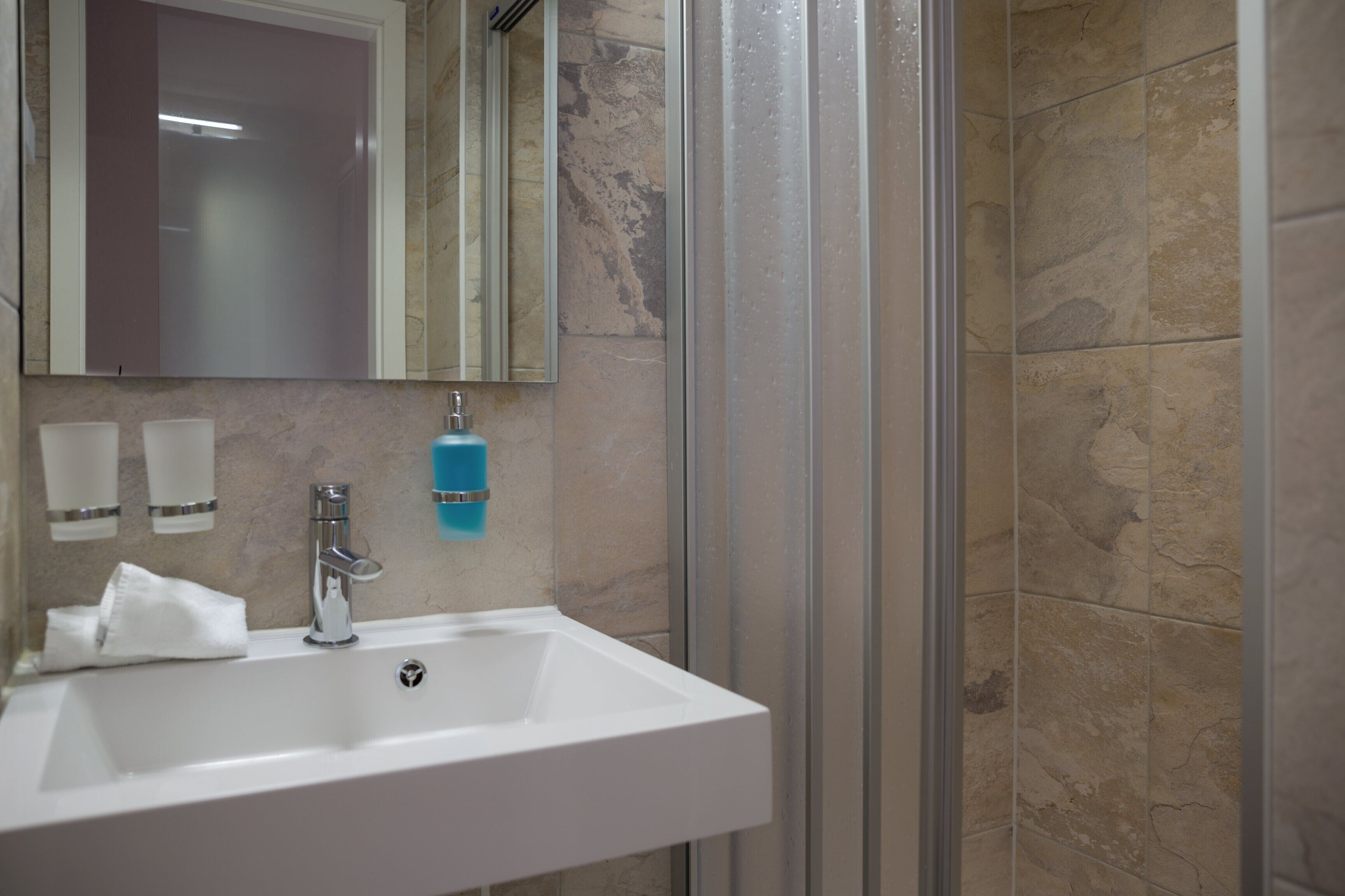 Une salle de bain avec un lavabo blanc, un miroir, un distributeur de savon bleu et une paroi de douche en verre.