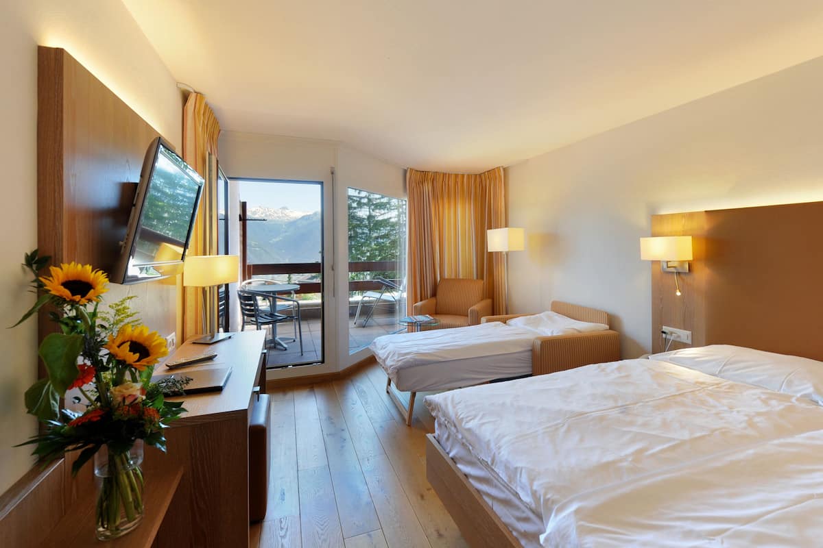 A hotel room with two beds, a desk with a laptop, a vase of flowers, and a view of mountains through the window.