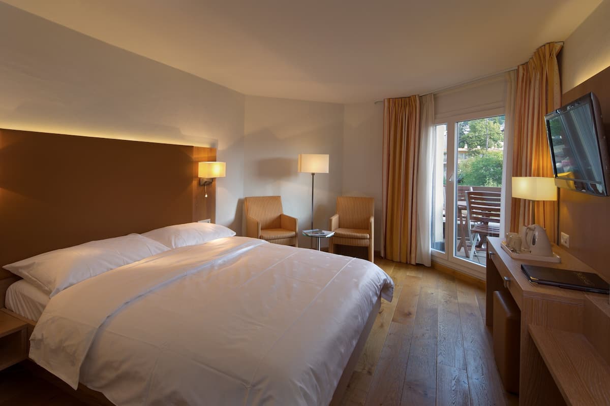 A hotel room with a large bed, two chairs, a desk with a television, and a window with curtains.