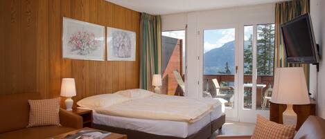 Habitación de hotel con cama, sofá, mesa de centro y televisión. Dos ventanas con cortinas y un balcón con vistas a montañas.