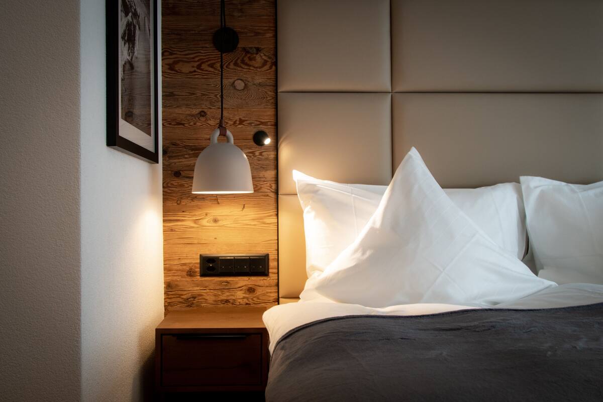 A neatly made bed with white linens, a wooden headboard, a wall-mounted light fixture, and a small bedside table.