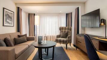 A modern hotel room with a sofa, armchair, desk, and TV.