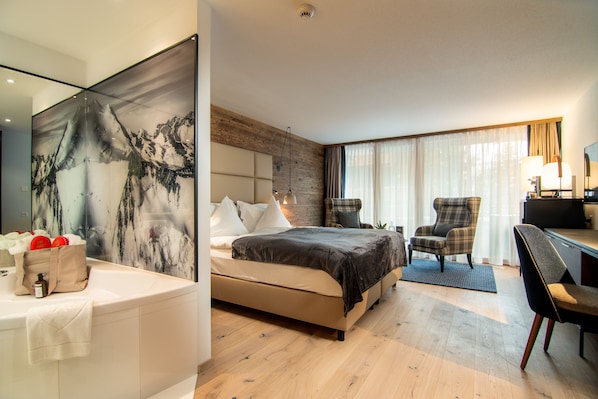 A modern hotel room with a large bed, a desk, a chair, and a bathroom with a large mirror and a painting. - Walliserhof Grand-Hotel & Spa Saas-Fee (Saas-Fee)