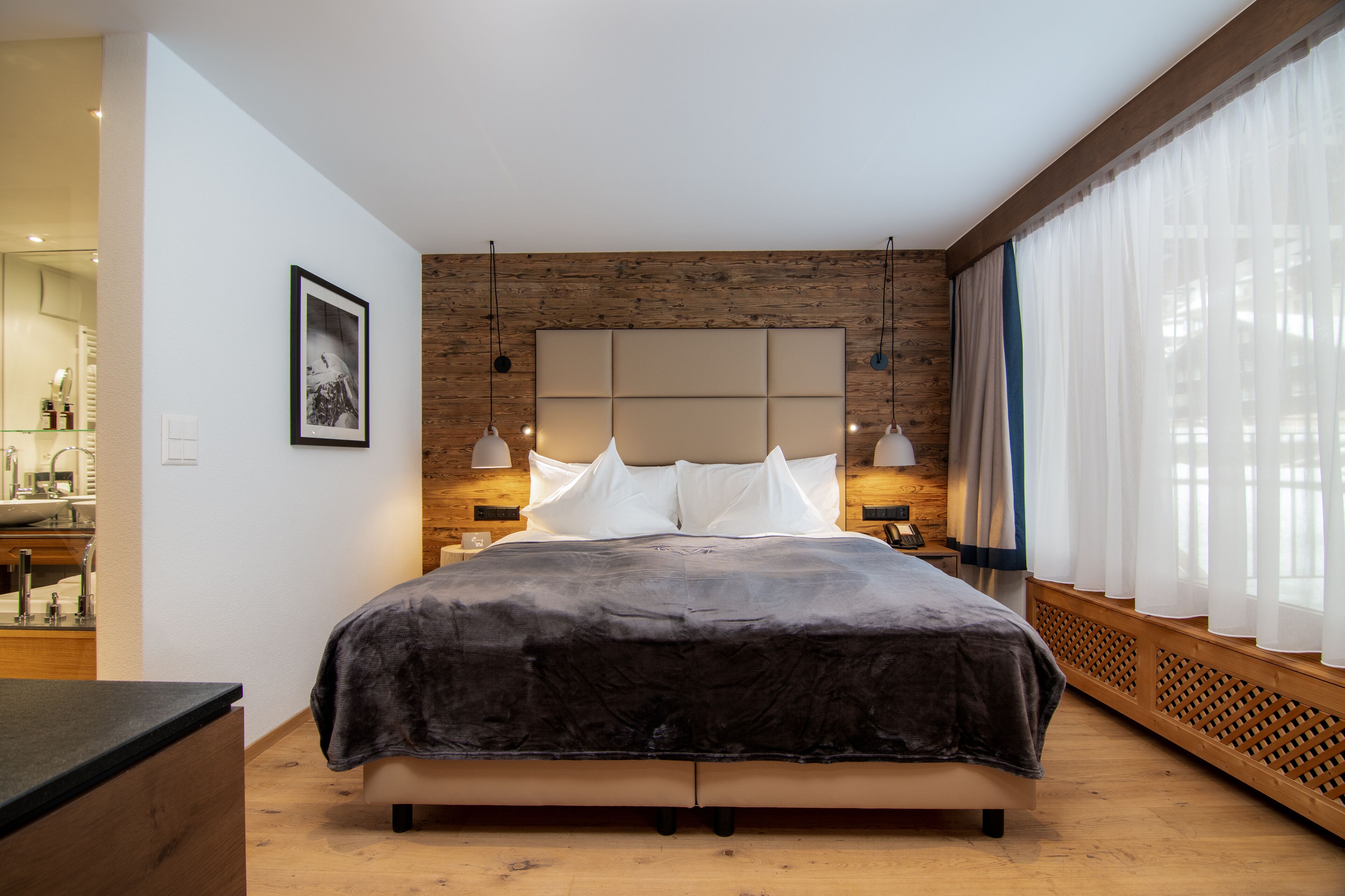 A modern bedroom with a large bed, wooden headboard, pendant lights, and a view of the city through sheer curtains.