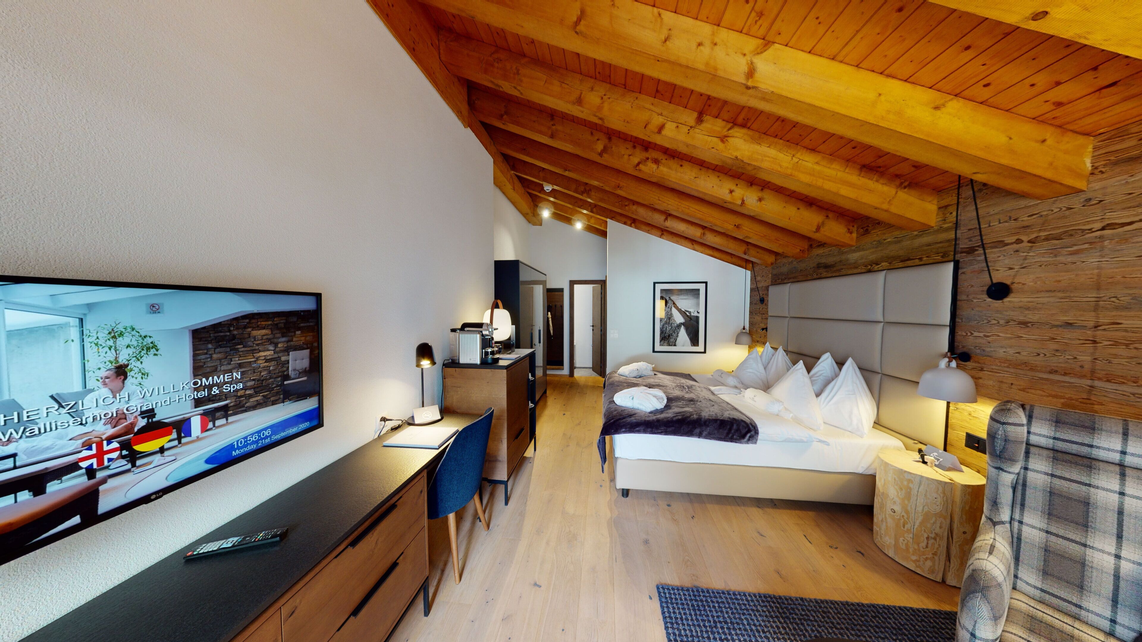 A modern hotel room with a wooden ceiling, a bed with white pillows, a desk with a TV, and a small bedside table.