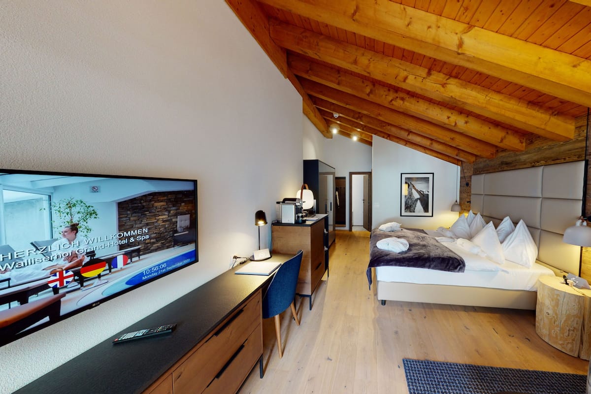 A modern hotel room with a wooden ceiling, a bed with white pillows, a desk with a TV, and a small bedside table.