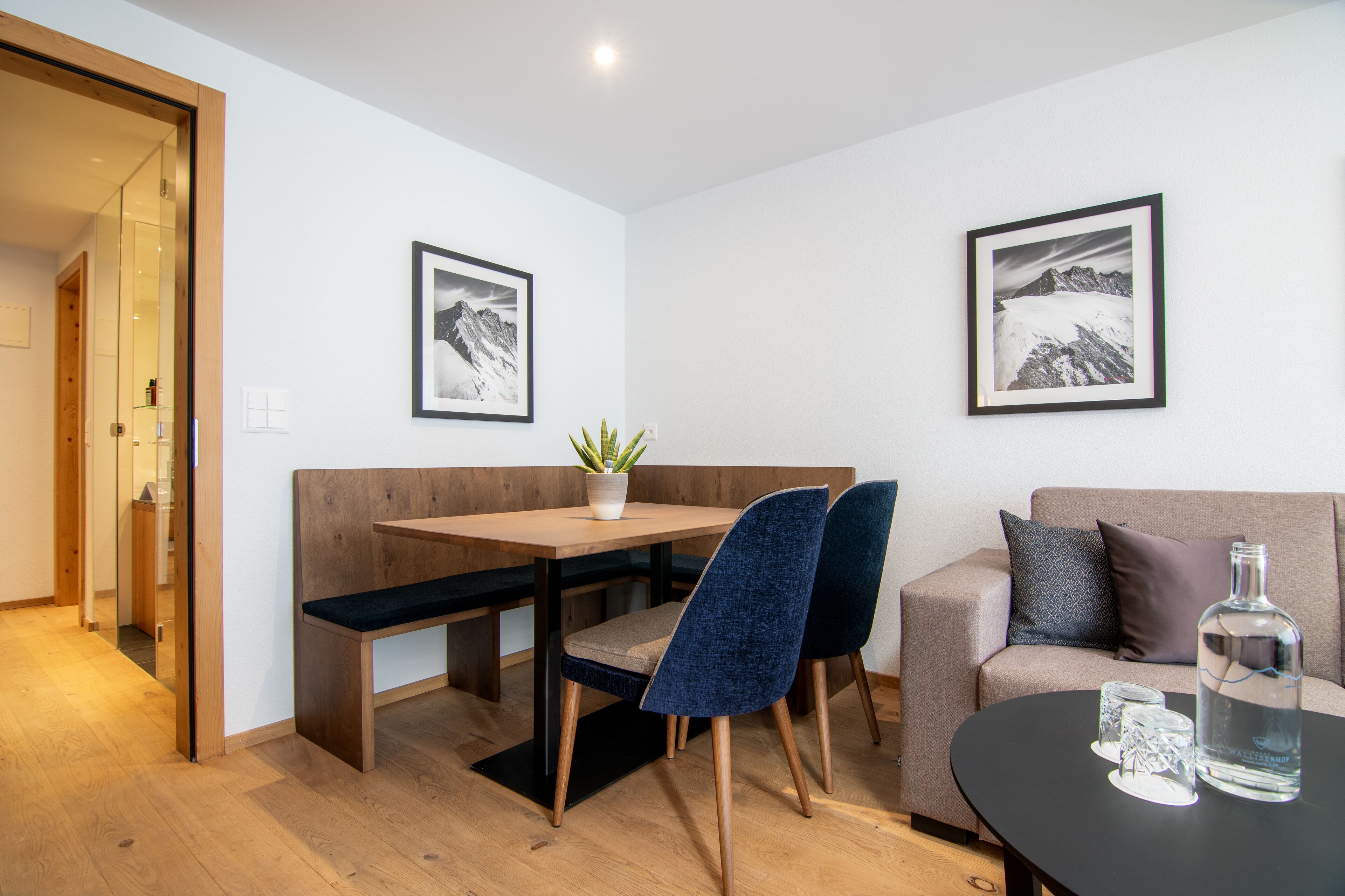 Un salón moderno con un banco de madera, dos sillas azules, una mesa de vidrio y dos cuadros en la pared.
