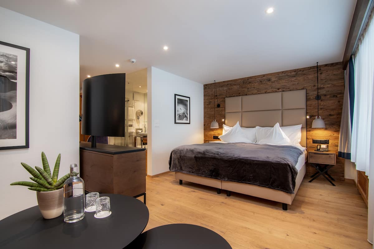 A modern hotel room with a large bed, a flat-screen TV, a wooden accent wall, and a small table with a plant and glasses.