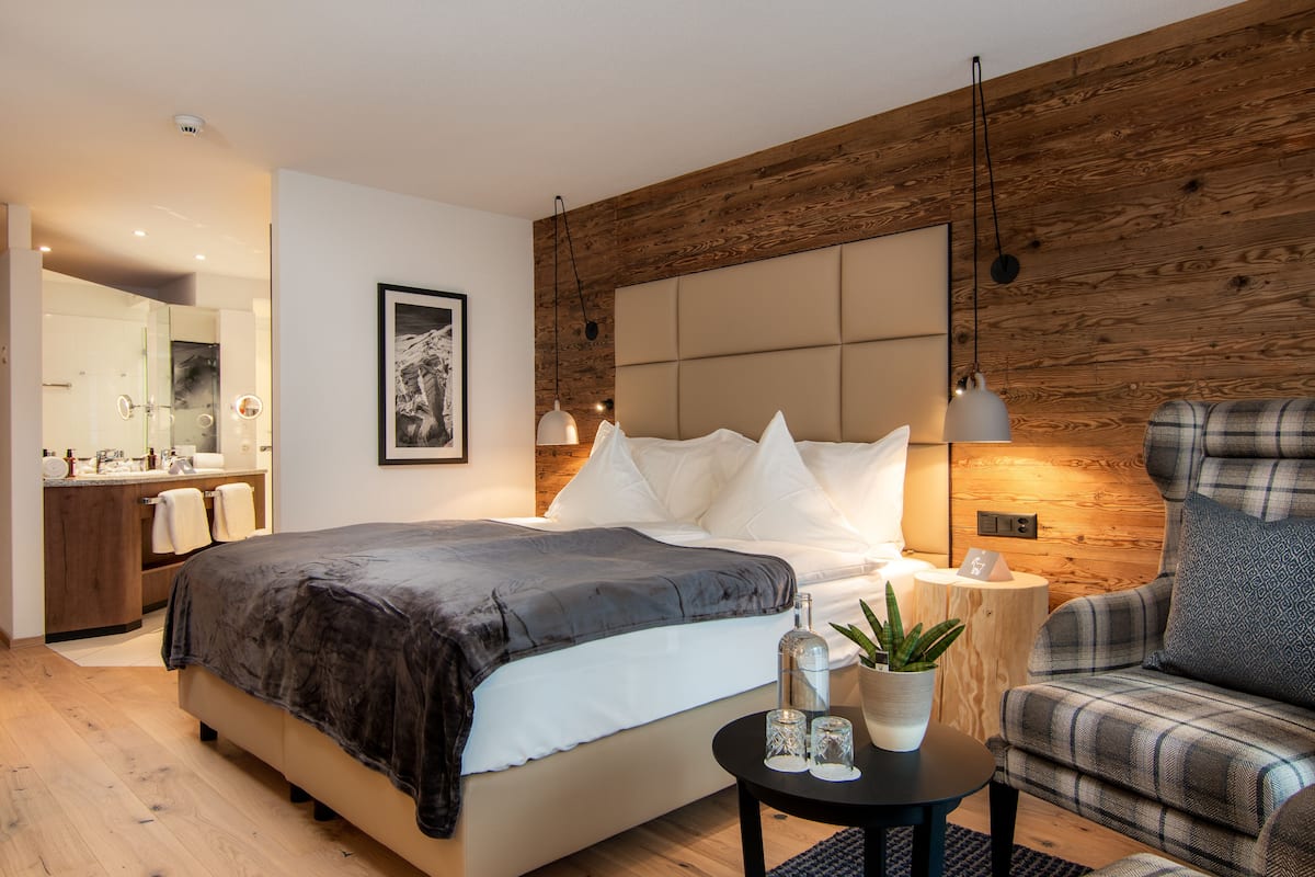 A modern bedroom with a large bed, a wooden accent wall, a bedside table with a plant, and a plaid armchair.