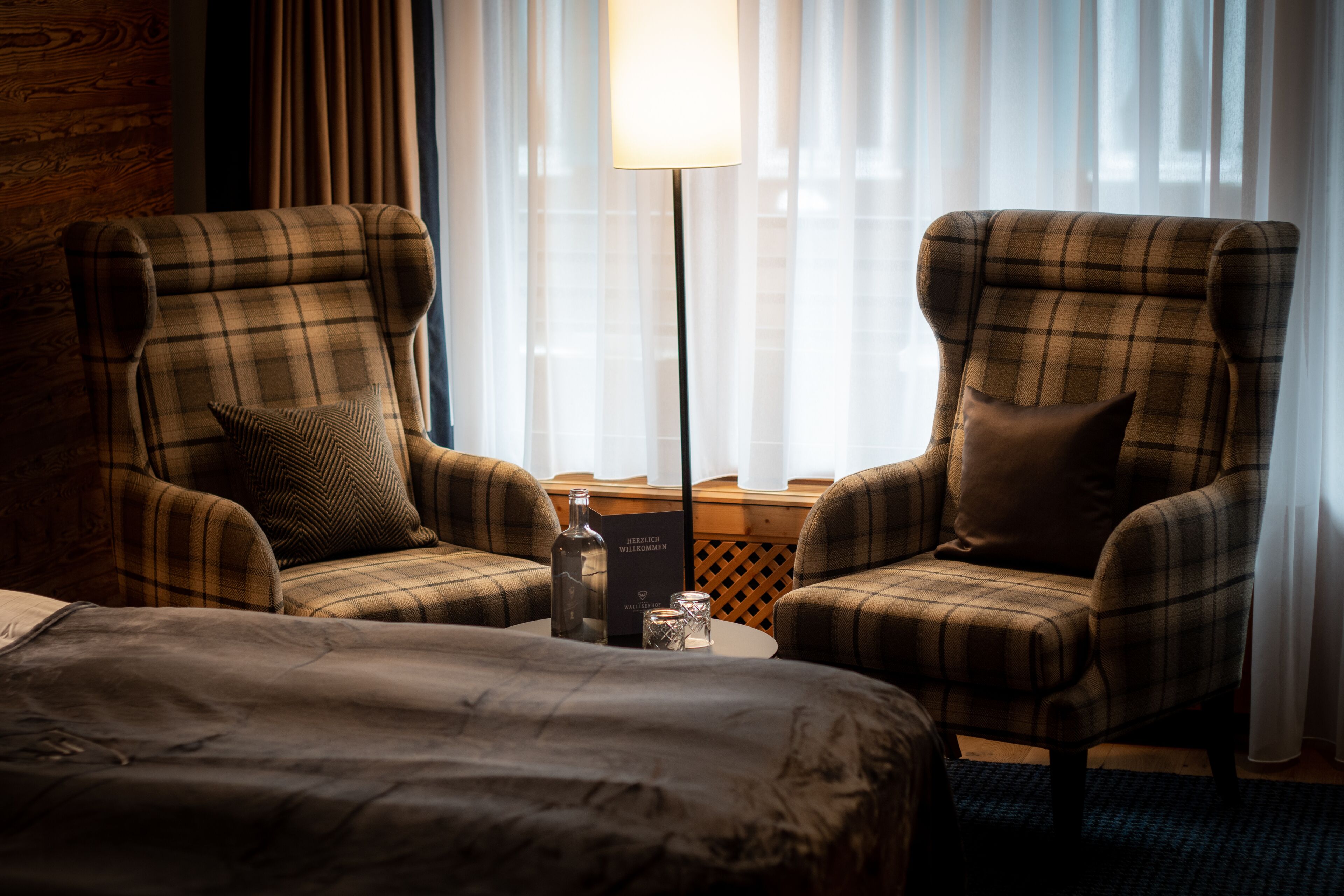 A hotel room with two plaid armchairs, a small table with a bottle and glasses, and a bed.