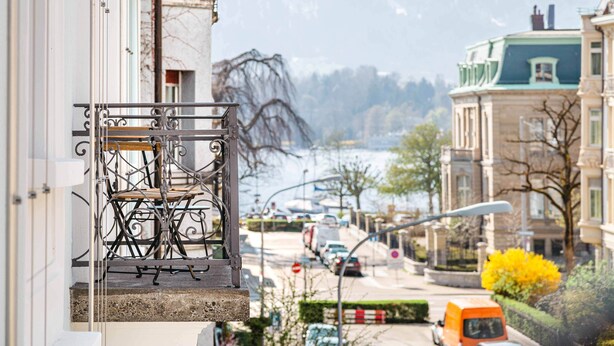 Un balcone con ringhiera in ferro battuto e una sedia, affacciato su una strada con auto parcheggiate e edifici sullo sfondo.