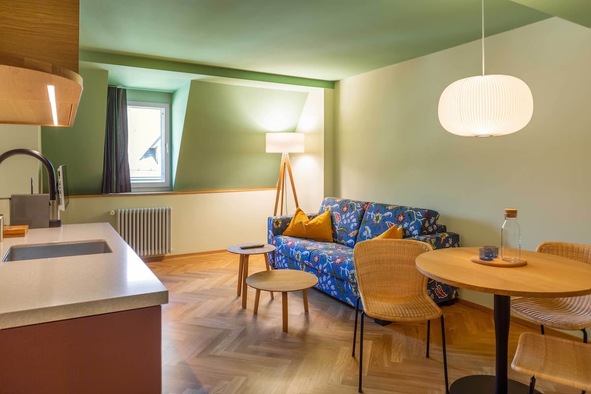 A modern kitchen with a central island, a dining area with a round table and chairs, a sofa with colorful cushions, and a floor lamp.