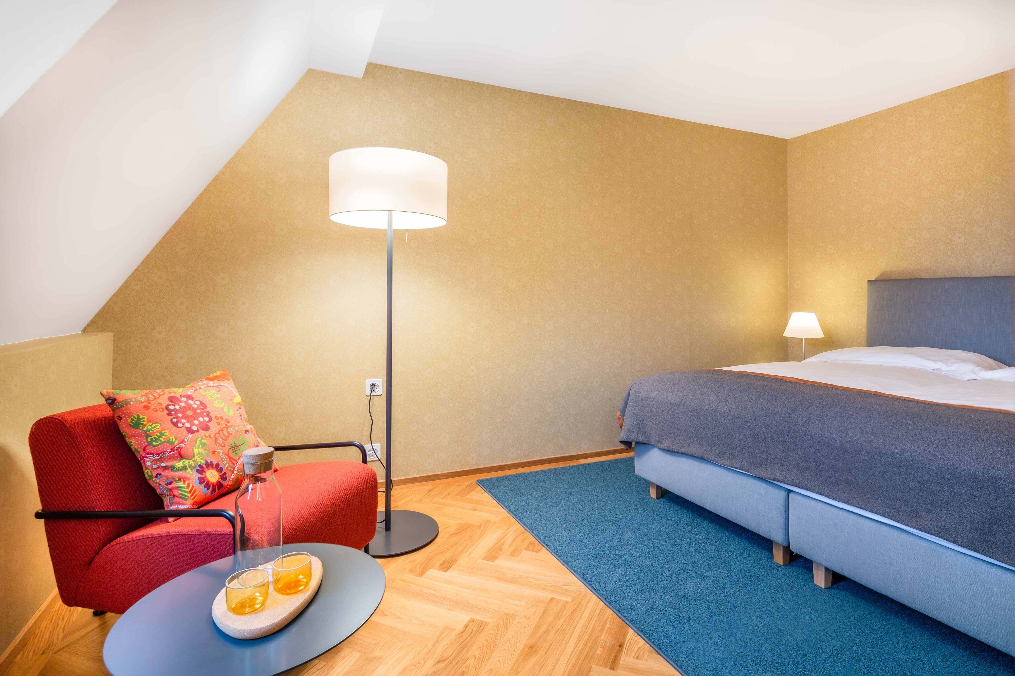 A hotel room with a bed, a red armchair, a floor lamp, and a small table with a tray of orange juice.
