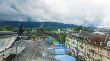 A rooftop terrace with blue folding chairs and potted plants, overlooking a cityscape with historic buildings and a river.