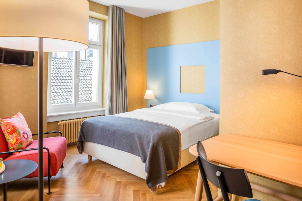 A hotel room with a bed, a desk, a chair, a lamp, a TV, and a window with curtains.