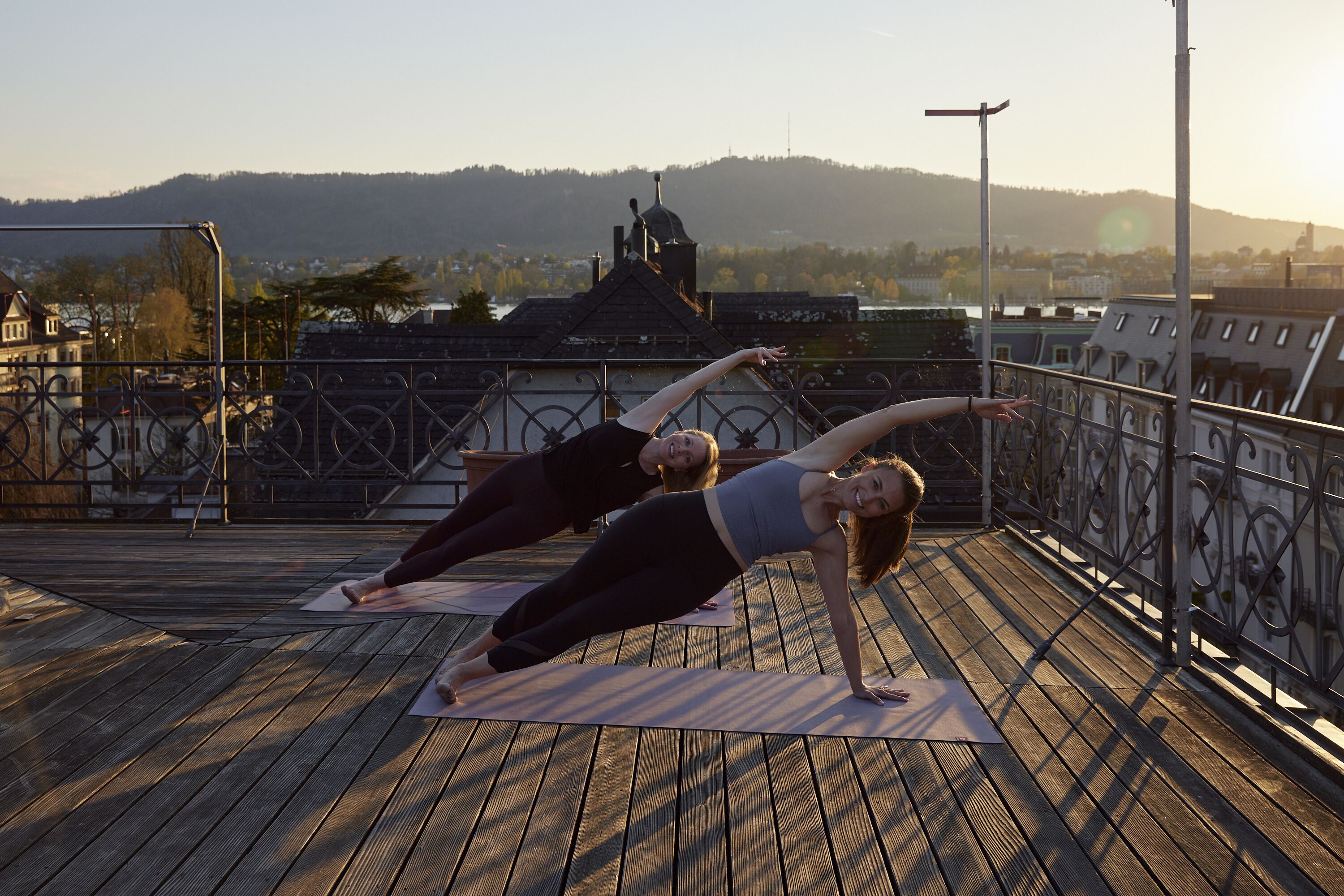 Zwei Personen praktizieren Yoga auf einer Holzterrasse, im Hintergrund eine Stadtlandschaft und Hügel.