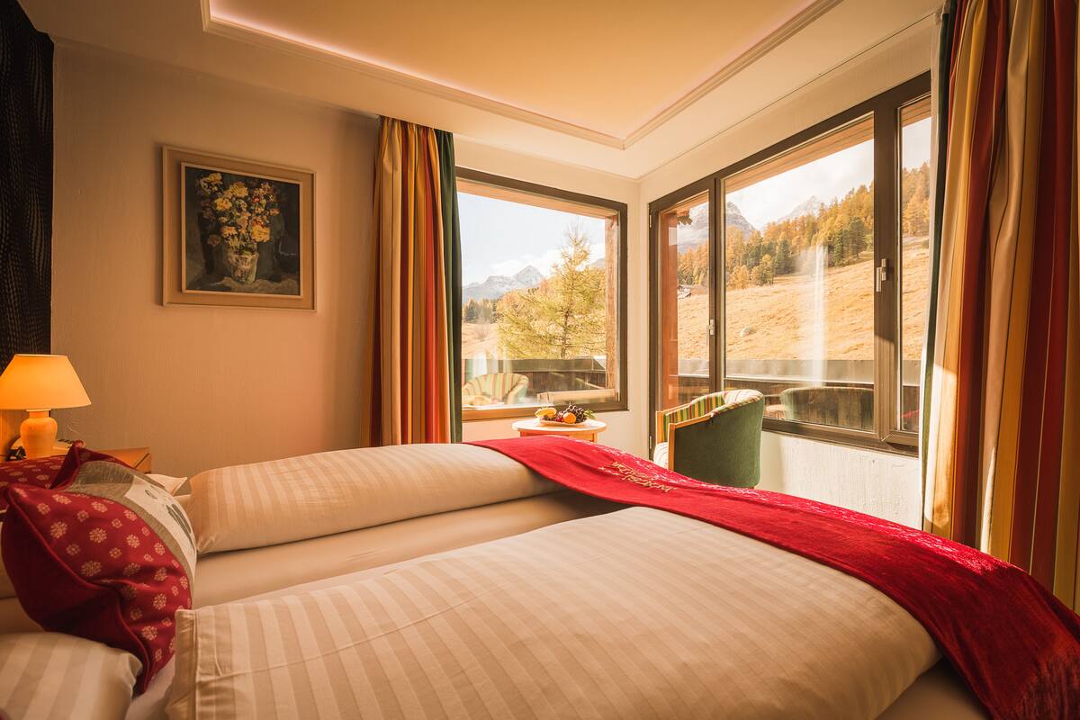 A hotel room with two beds, a window with a mountain view, a painting on the wall, and a lamp on the nightstand.