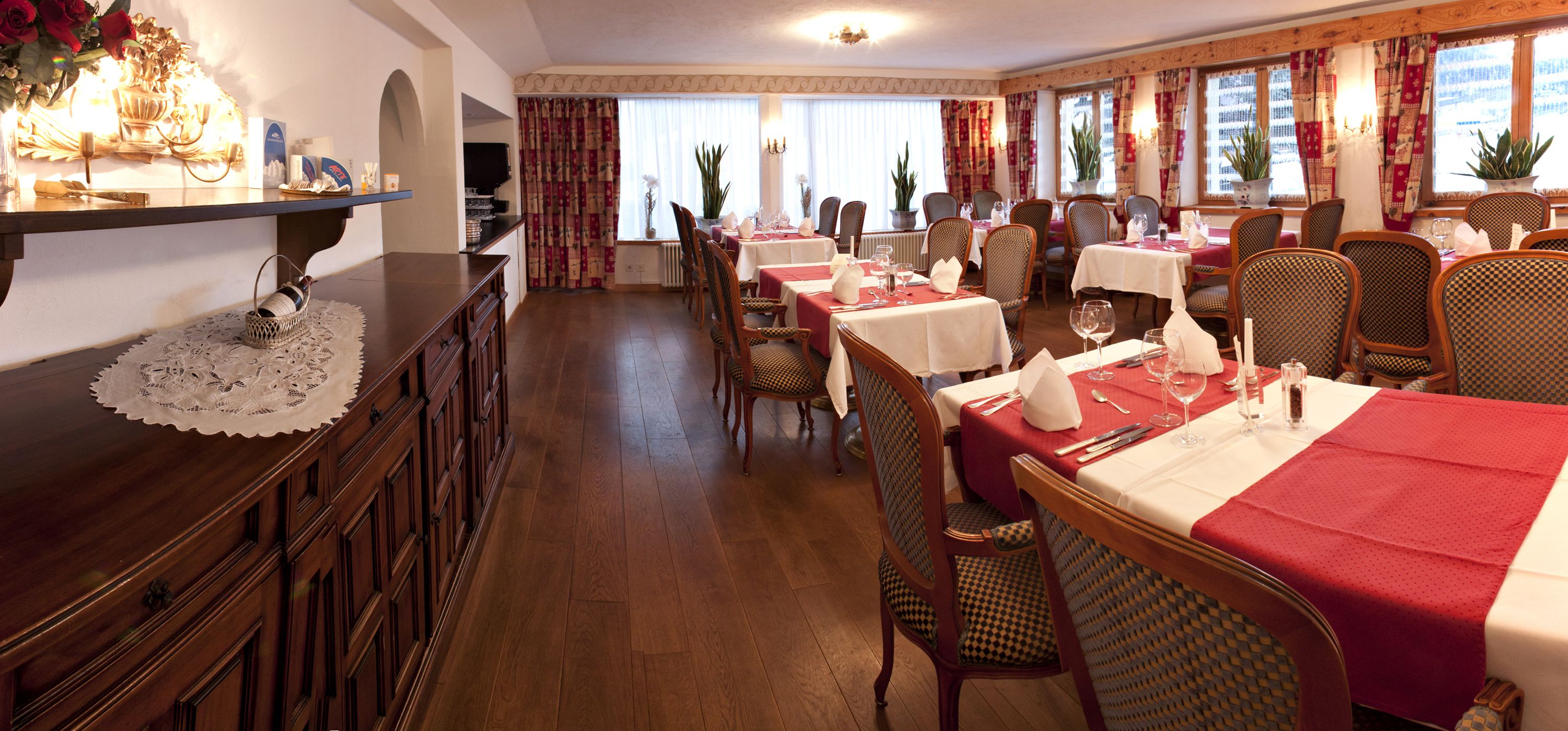 Un restaurant doté de nombreuses tables, chacune recouverte d’une nappe blanche et agrémentée de serviettes rouges. On y trouve des chaises en osier et un comptoir en bois surmonté d’une chemin de table en dentelle.