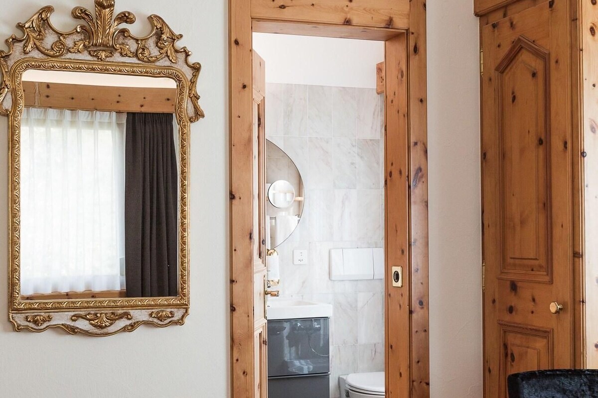 Una habitación con una puerta de madera, un espejo con un marco decorativo, una colcha roja y una silla roja.