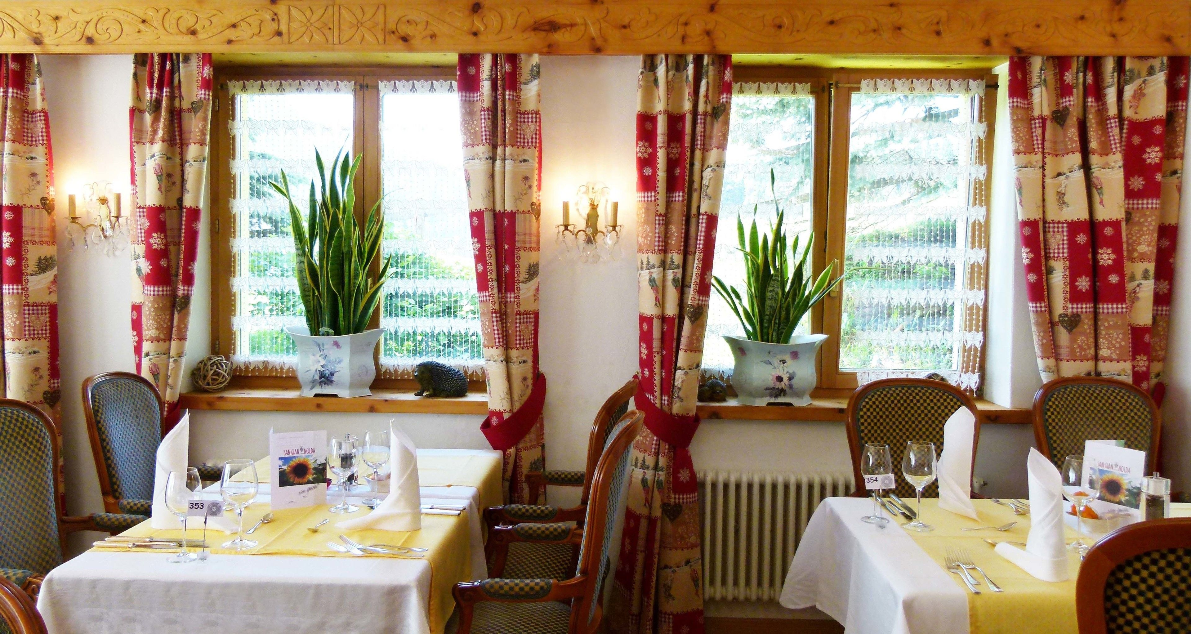 Un comedor con mesas preparadas para dos, cada una con un número, copas de vino y cubiertos. Hay plantas en macetas en el alféizar de la ventana y el techo es de madera rústica.