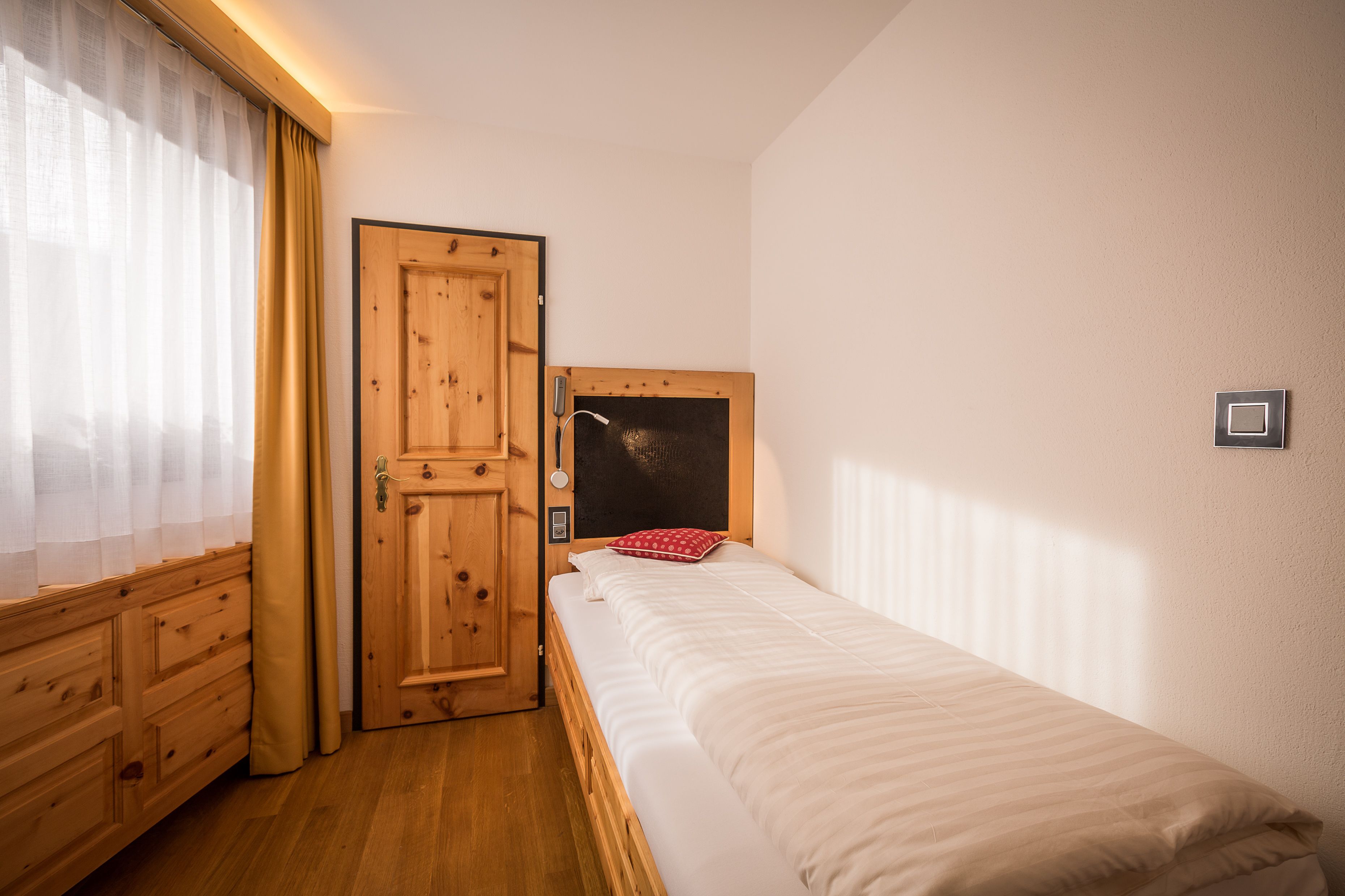 A small, neatly arranged bedroom with a wooden wardrobe, a single bed, and a window with sheer curtains.