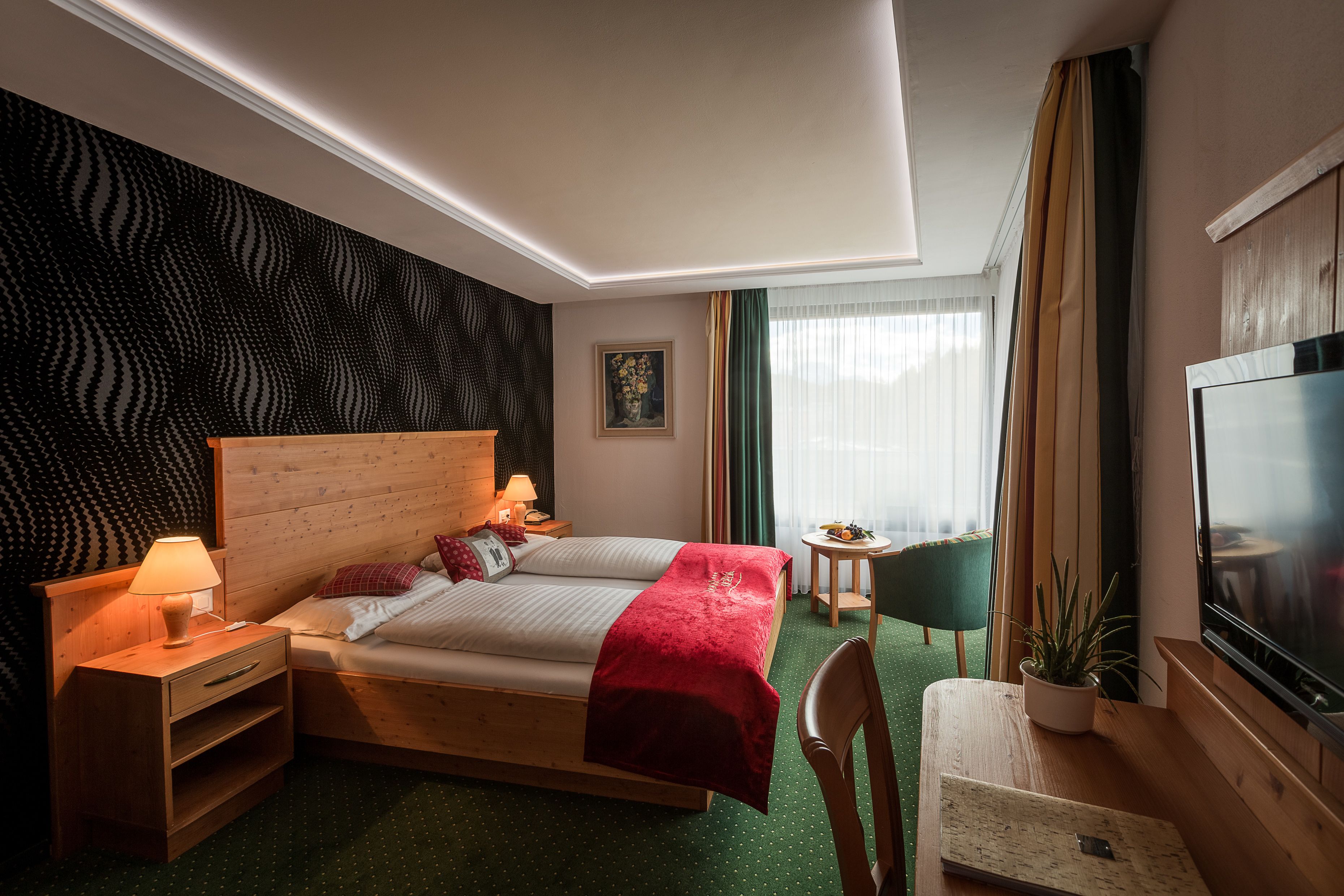 A hotel room with a wooden bed, a bedside table with a lamp, a small table with a plant, a chair, and a TV.