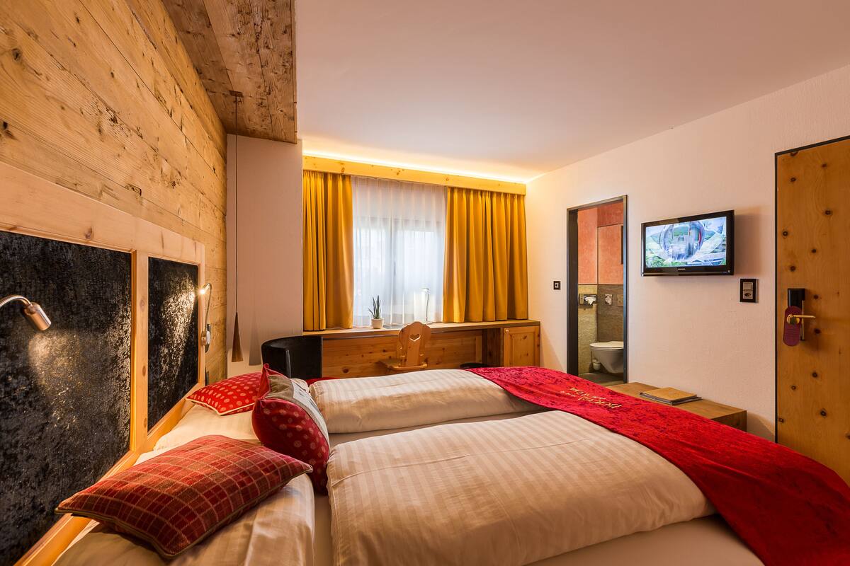 A hotel room with a wooden ceiling, a bed with red and white bedding, a wooden dresser, a television, and a bathroom visible through an open door.