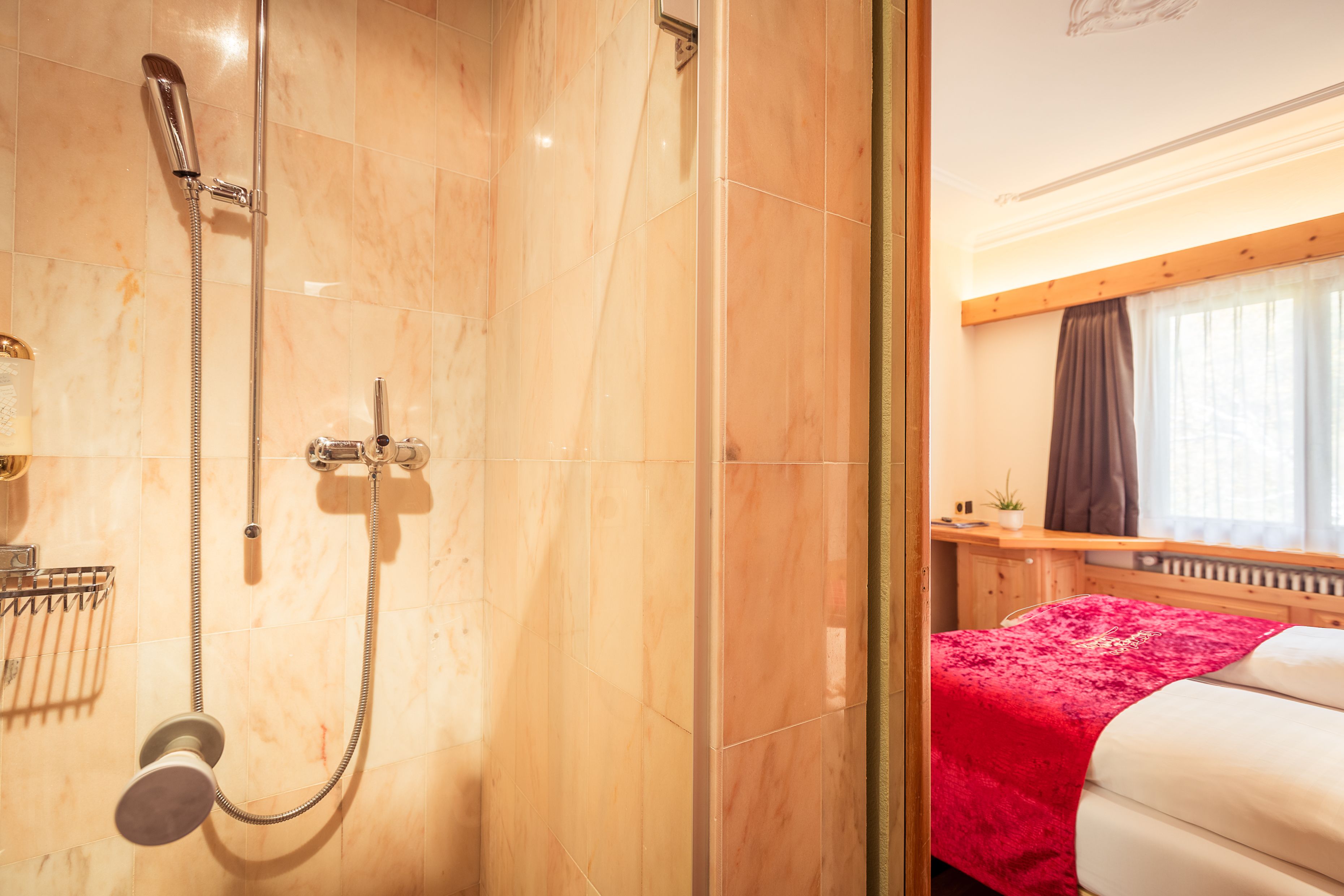 Un baño de hotel con ducha, una cama con una manta roja y un escritorio de madera con una planta.