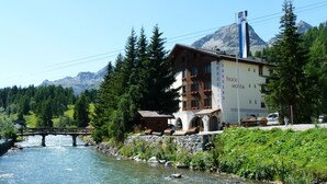 Un hotel con el paisaje de montañas al fondo, un río en primer plano y un puente de madera.