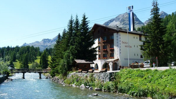 A hotel with a mountain backdrop, a river in front, and a wooden bridge.