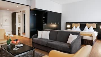 A modern hotel room with a grey sofa, a yellow armchair, a dining table with fruit, a bed with white bedding, and a dark wardrobe.