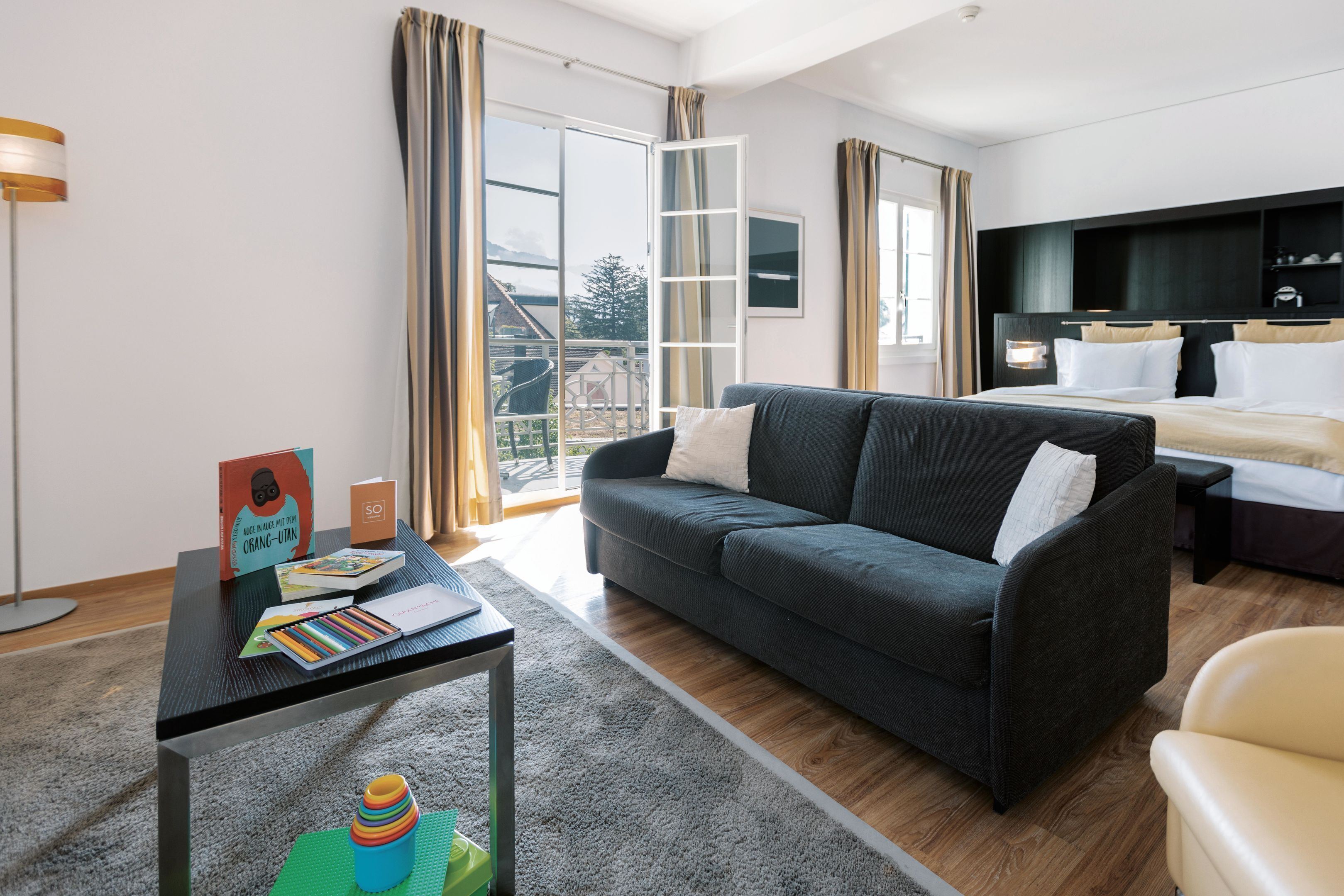 Una habitación de hotel moderna con un sofá gris oscuro, una mesita con libros y un juguete, una cama con ropa de cama blanca y un ventanal con cortinas.