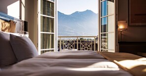 A hotel room with a bed, a balcony with a view of mountains, and a window with curtains.