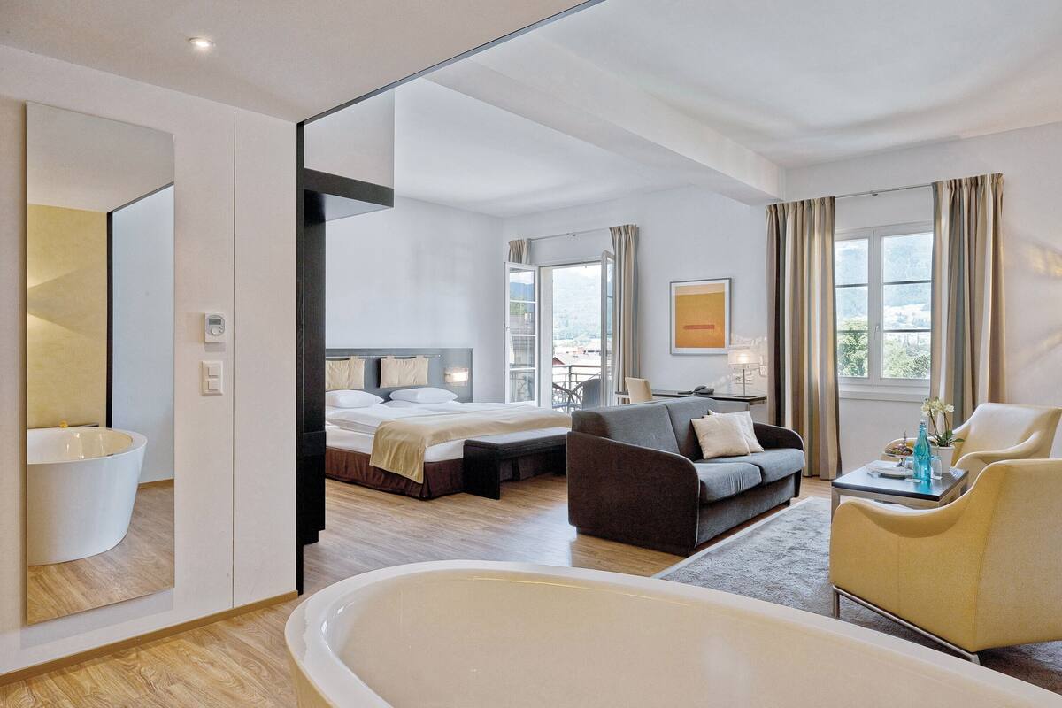 A modern hotel room with a large bed, a sitting area with a sofa and armchairs, a bathtub, and a view of the city.