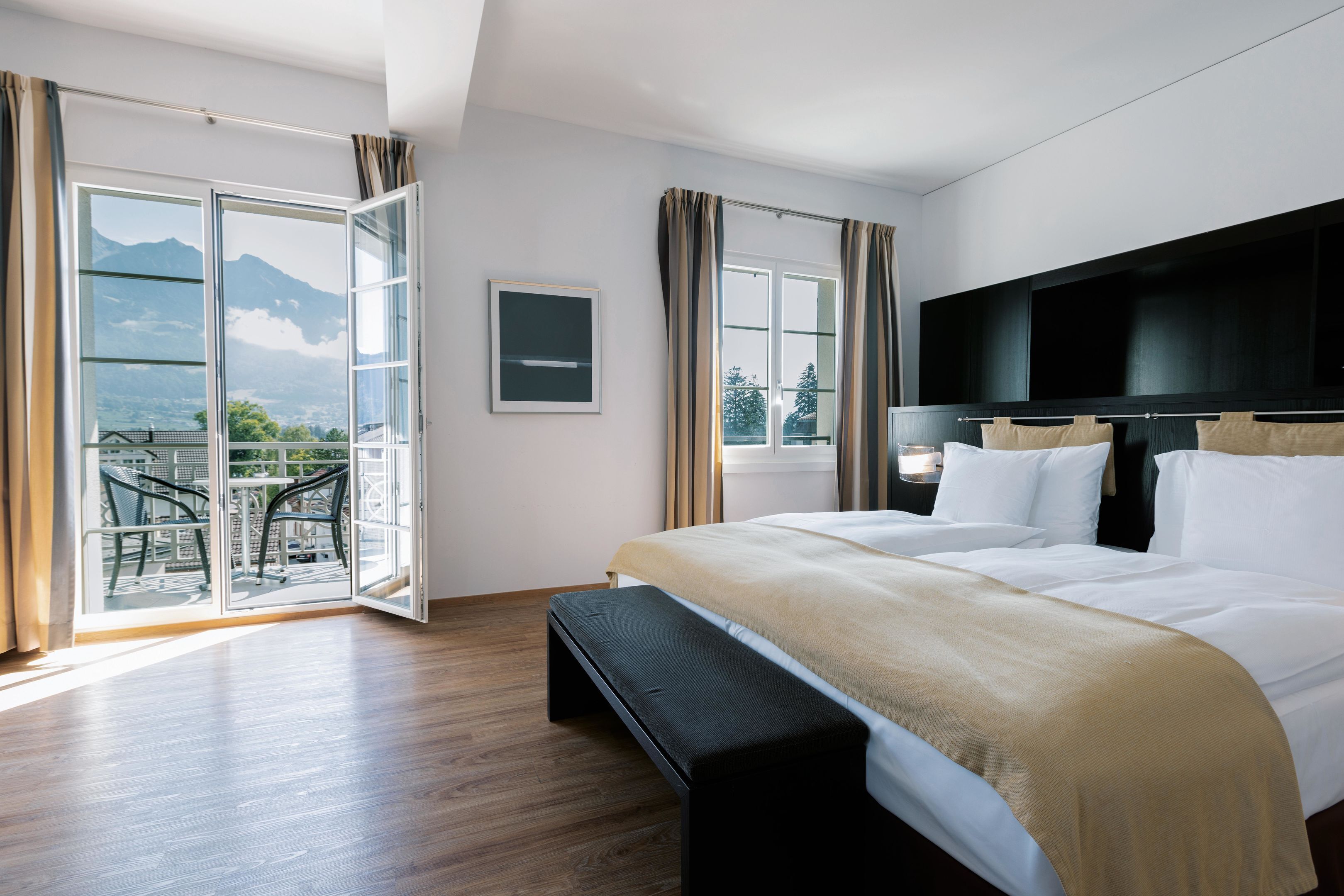 Quarto com cama, um banquinho, televisão e varanda com vista para as montanhas.