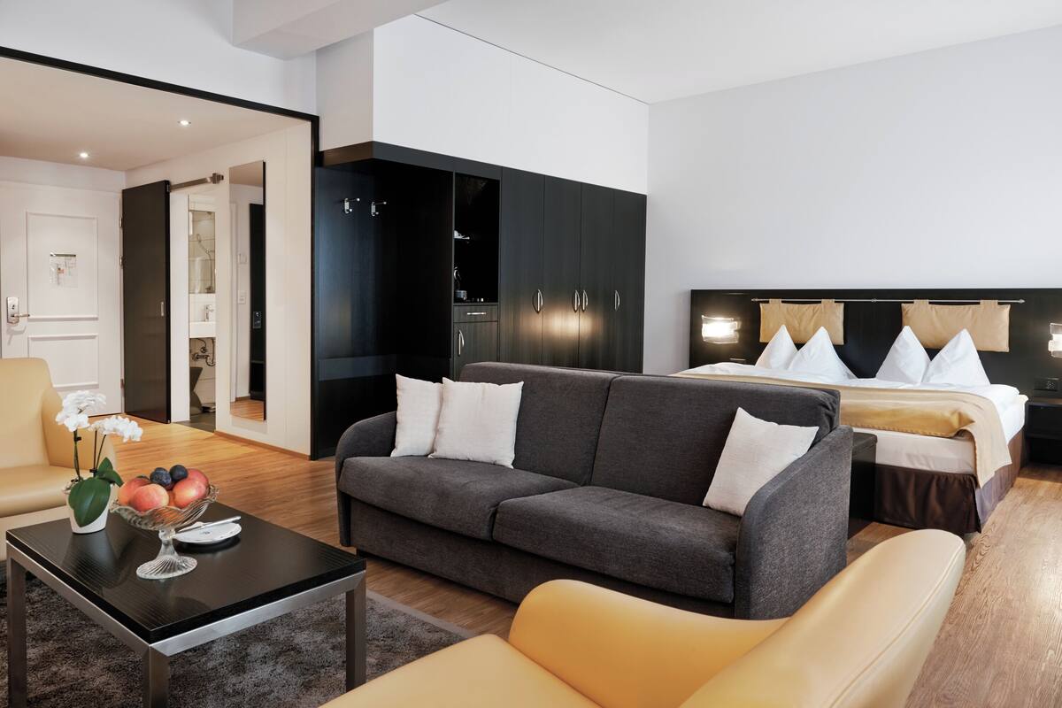 A modern hotel room with a grey sofa, a yellow armchair, a dining table with fruit, a bed with white and beige bedding, and a black wardrobe.