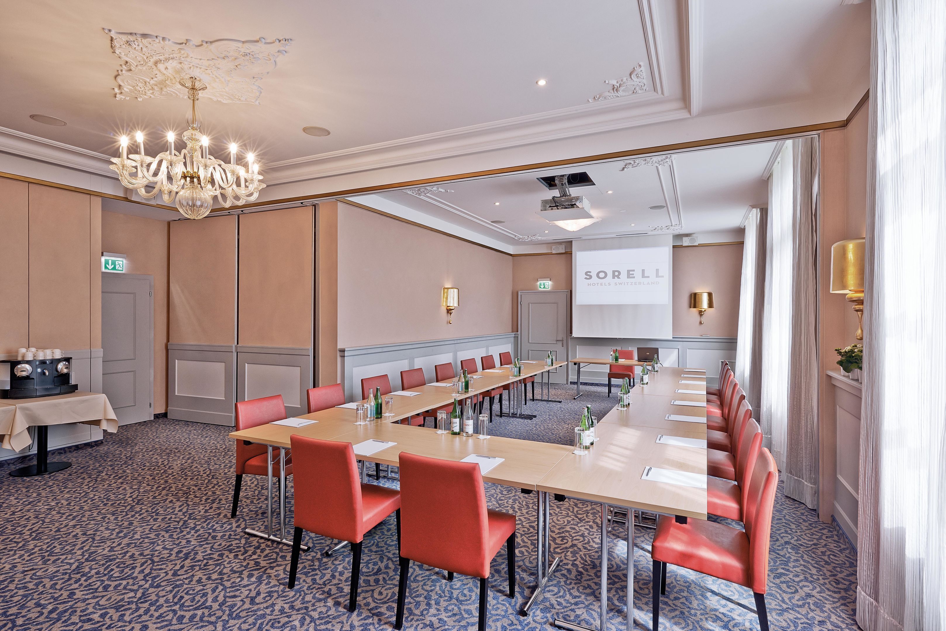 A conference room with a long table, red chairs, a chandelier, and a projector screen.