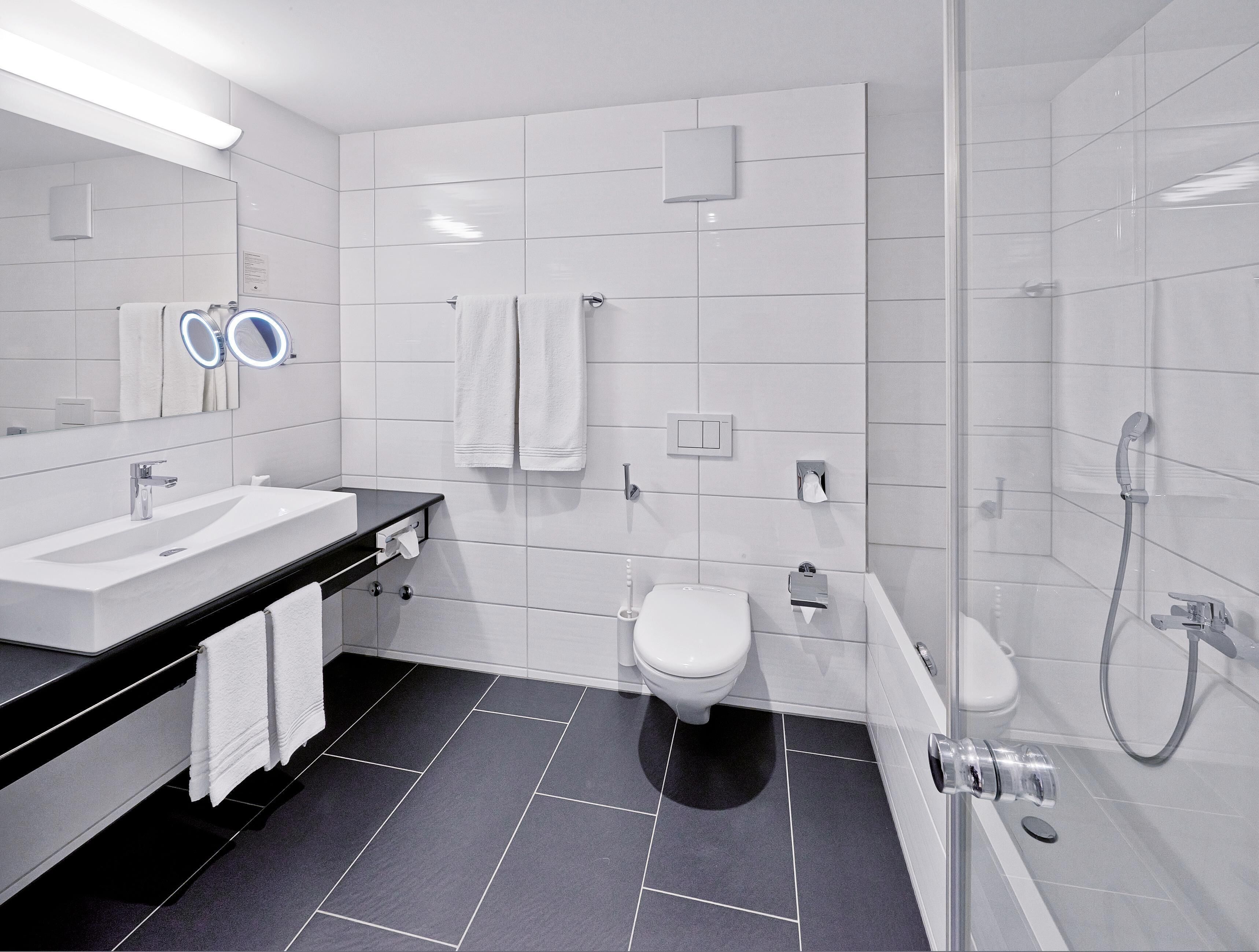A modern bathroom with a large mirror, white tiles, a black countertop, a toilet, a bidet, and a glass-enclosed shower area.