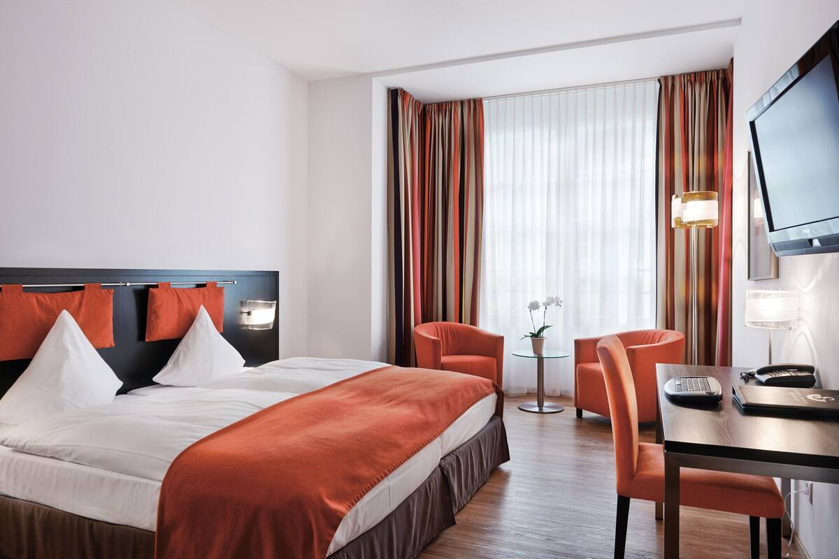 A hotel room with a large bed, a desk with a chair, a TV, and a window with curtains.